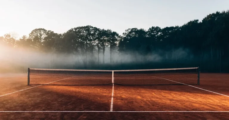 Ein Tennisplatz aus Sand bei Sonnenaufgang mit gespanntem Netz, Nebel zwischen den Bäumen am Rand